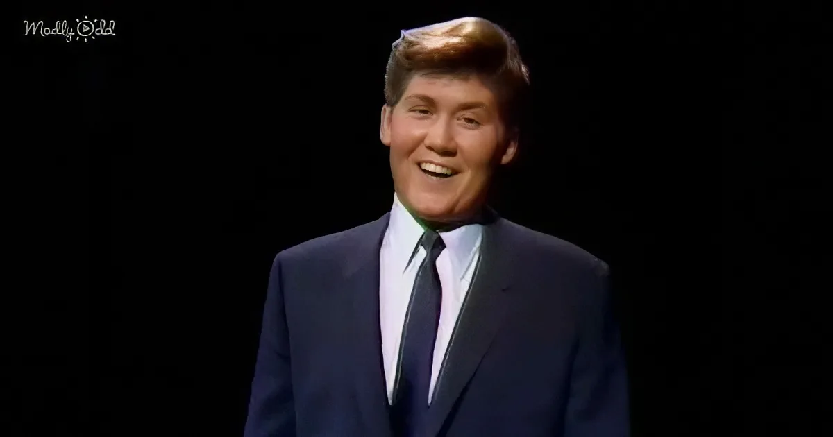 A man in a dark suit and tie smiles brightly against a black background.