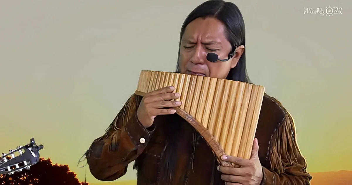 A person in a brown outfit plays a wooden pan flute, wearing a headset, with a sunset background.