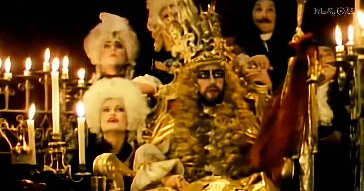 A man in ornate gold armor and crown sits on a throne, surrounded by people in period costumes and lit candles.