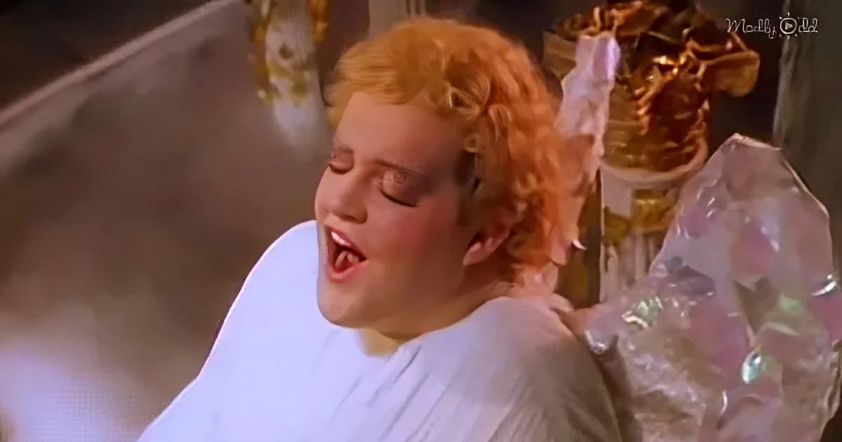 A person with curly red hair, wearing a white robe, is lying down with eyes closed, appearing to be in a relaxed or...