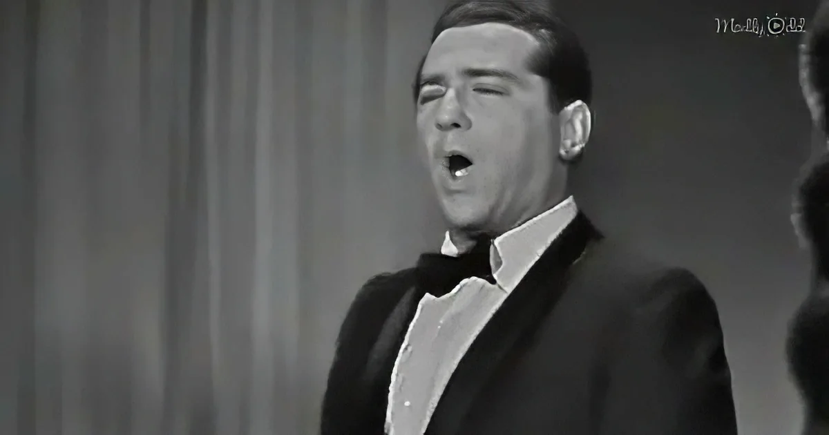 A man in a tuxedo sings passionately, mouth open, against a curtain backdrop.