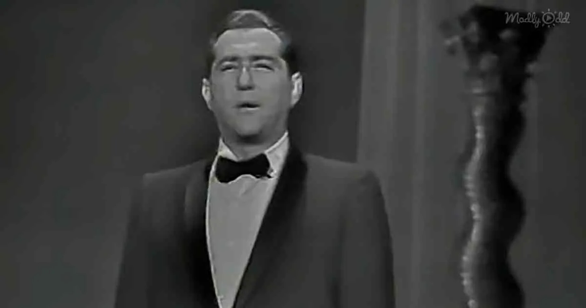 A man in a tuxedo sings on stage, mouth open, with a dark background and a trophy-like object to the side.