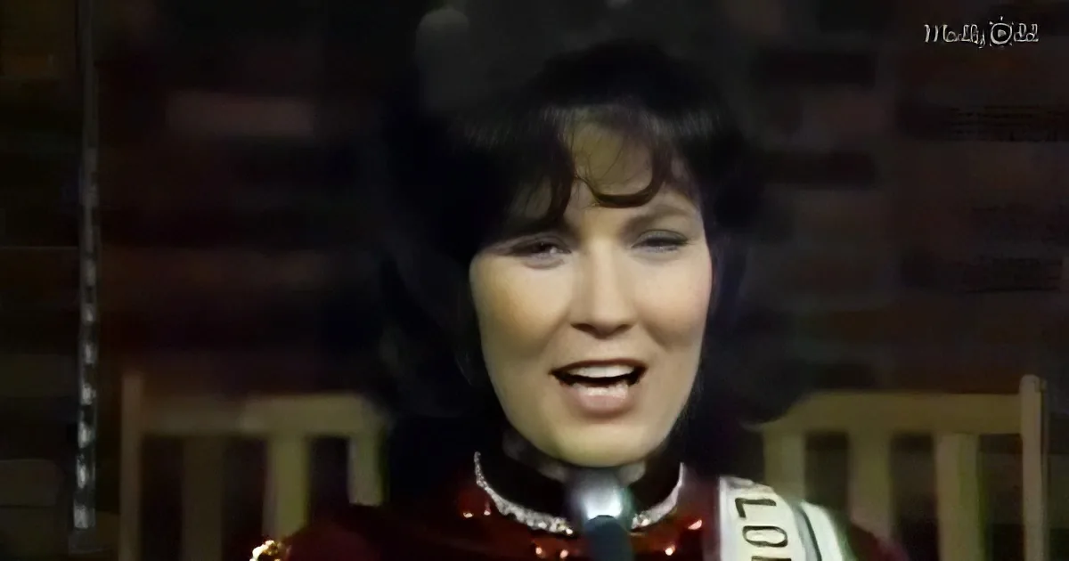 A person in a red outfit sings into a microphone, with a dark background and a chair visible.