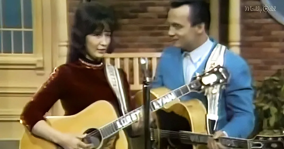 A woman in a red dress plays guitar while a man in a blue suit sings into a microphone.