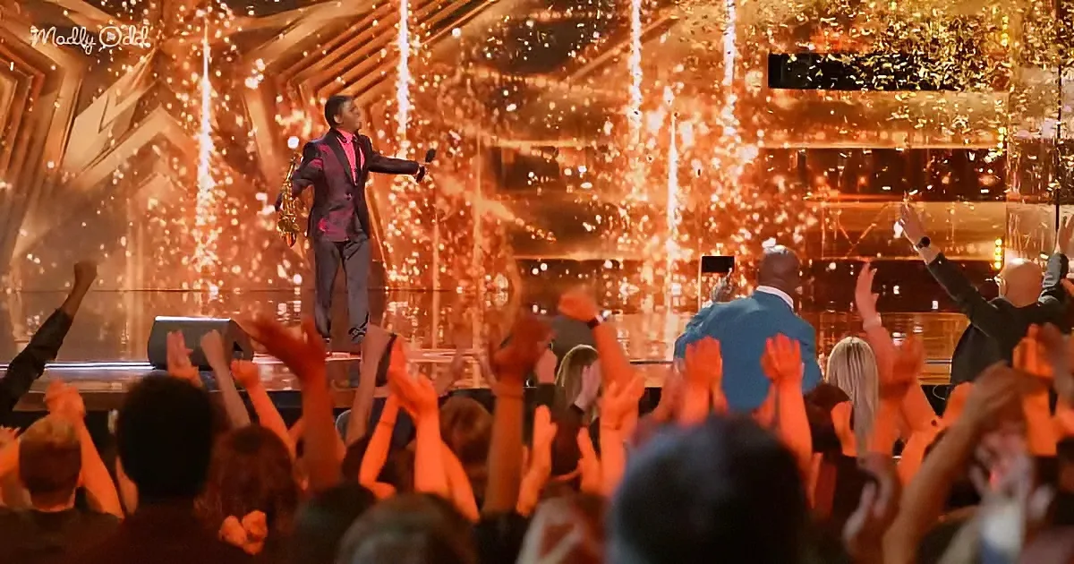 A man in a suit stands on stage, holding a microphone, surrounded by golden confetti and cheering crowd.