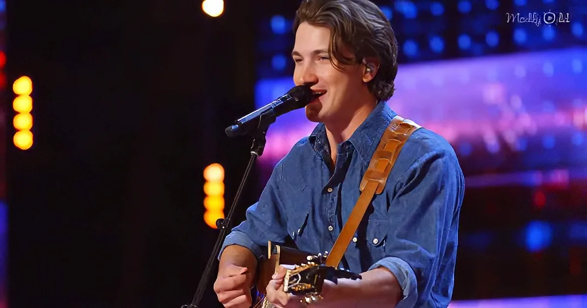 A man in a blue shirt sings into a microphone while playing a guitar on stage.