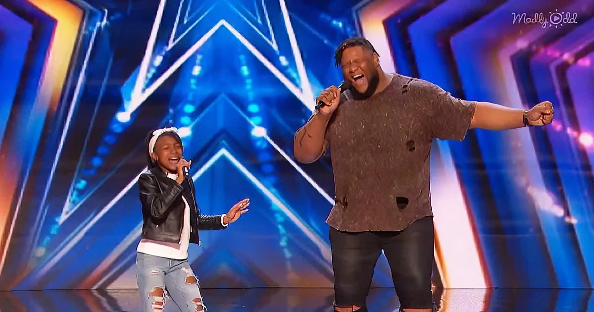 A man and a young girl sing passionately on a brightly lit stage with a large "A" backdrop.