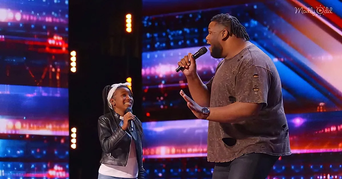 A man and a young girl sing on stage, both holding microphones, with colorful lights in the background.