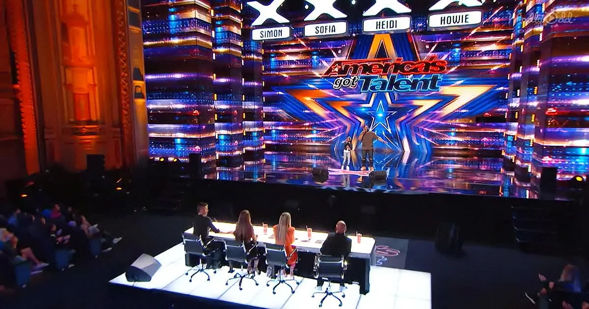 A performer stands on a brightly lit stage with judges seated at a table in front.
