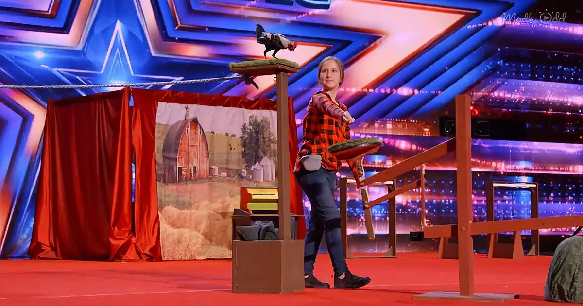 A woman in a plaid shirt stands on a stage with a chicken perched on a wooden structure.