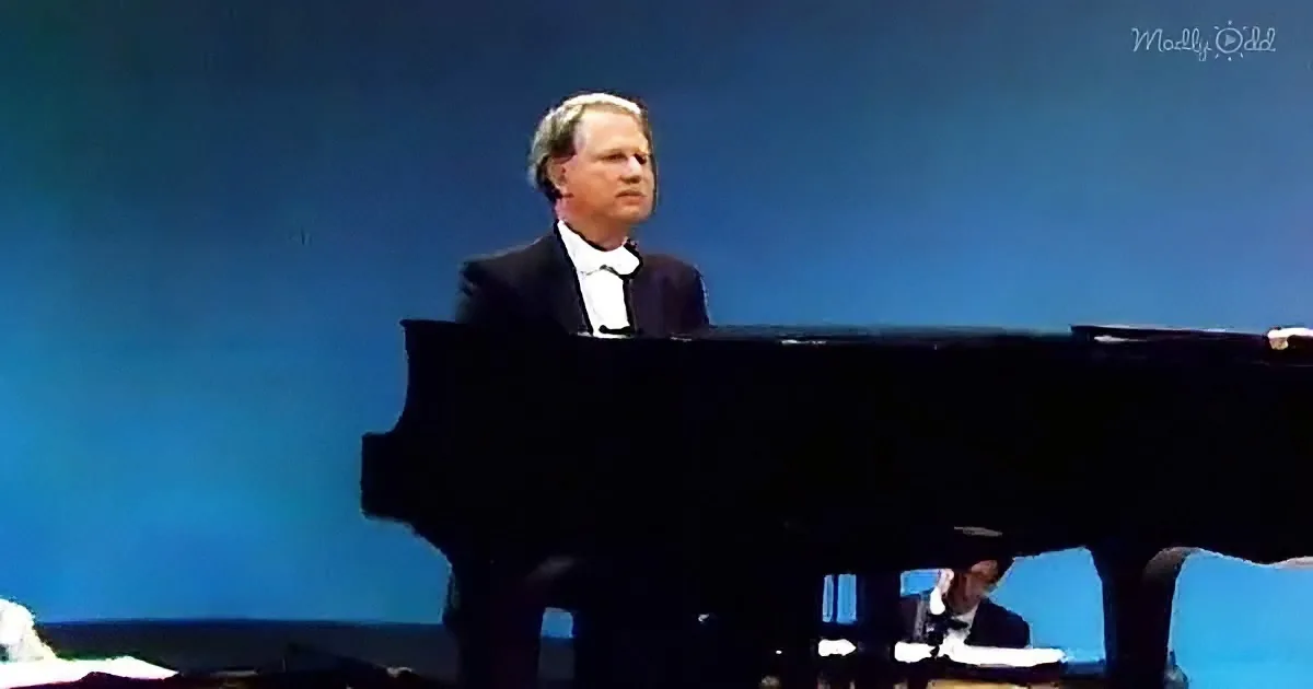 A man in formal attire plays a grand piano on stage, with a blue background.