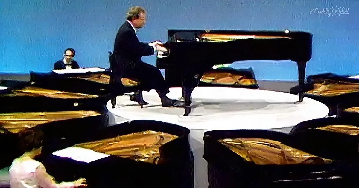 A man in a suit plays a grand piano on a stage with a blue backdrop.