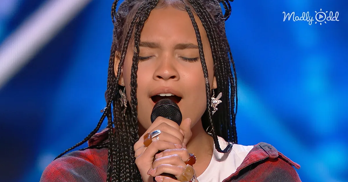 A person with braided hair sings into a microphone, wearing a red plaid jacket and earrings, against a blue background.