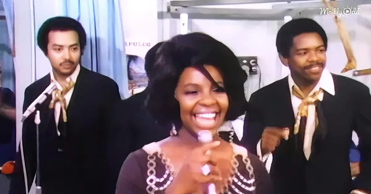 A smiling woman in a black and white outfit holds a microphone, flanked by two men in suits with gold scarves.