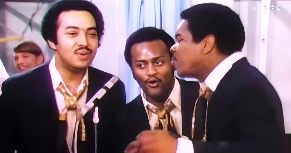 Three men in suits sing into microphones, one holding a glass, with a smiling background figure.