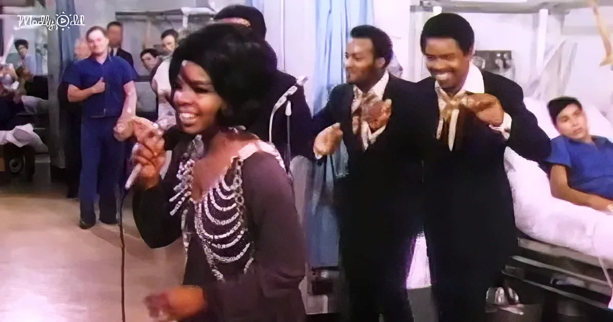 A woman in a skeleton costume sings into a microphone, surrounded by men in suits and nurses in blue scrubs.