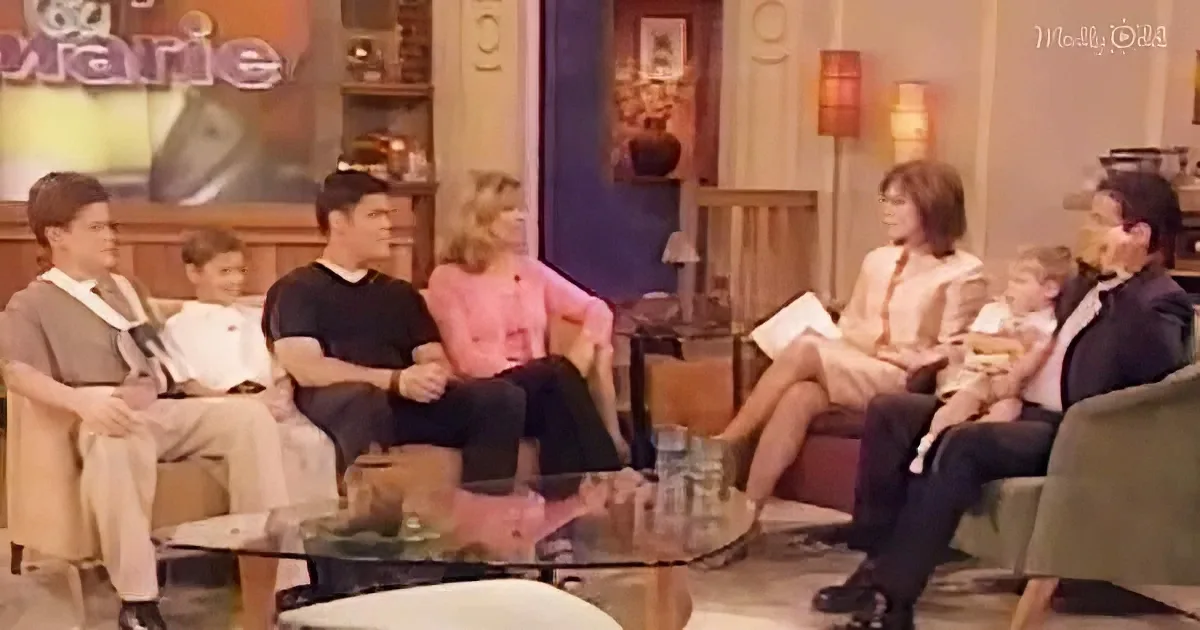 A group of people, including children and adults, are seated in a living room setting, engaged in conversation.