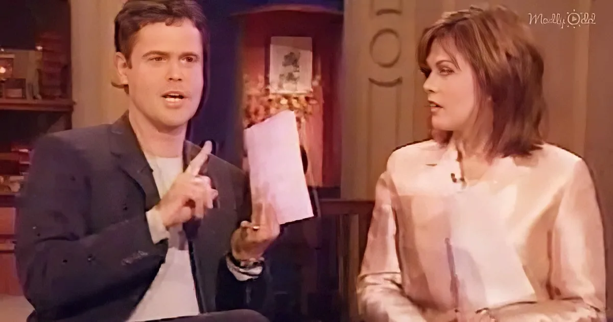 A man in a suit gestures while speaking, seated next to a woman in a light blouse on a talk show set.