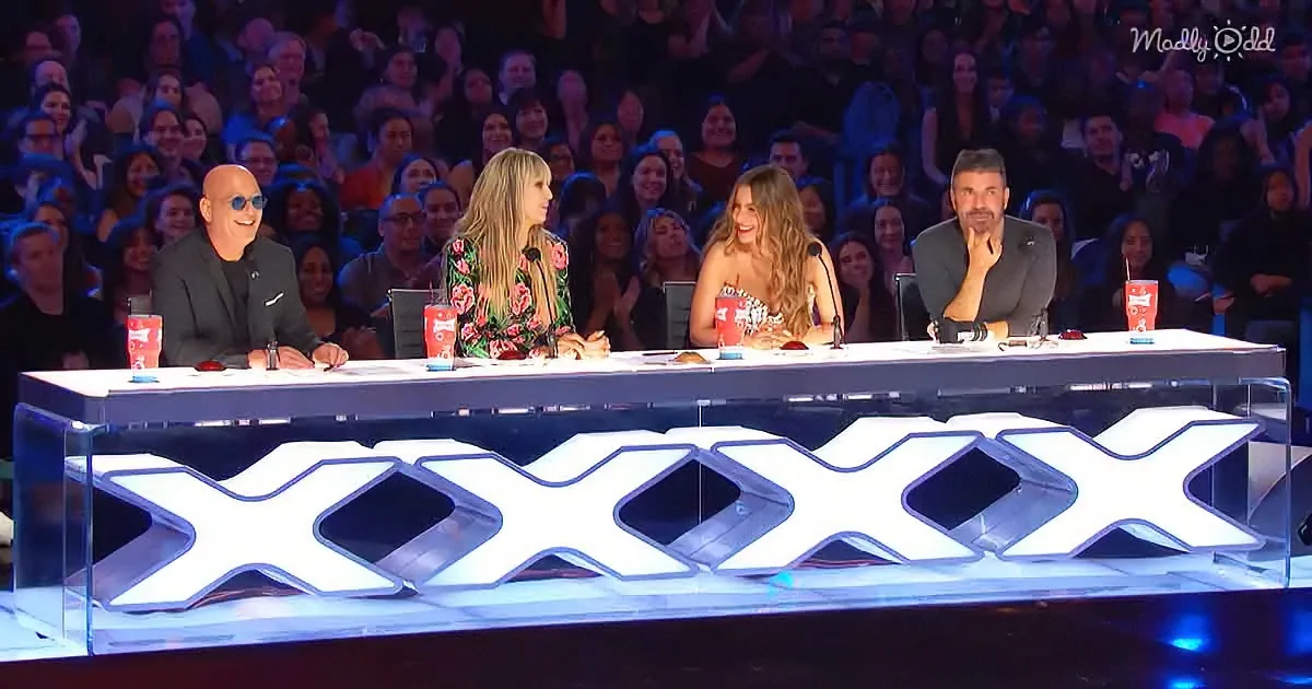 Four judges sit at a table with "XXXX" illuminated, in front of a cheering audience.