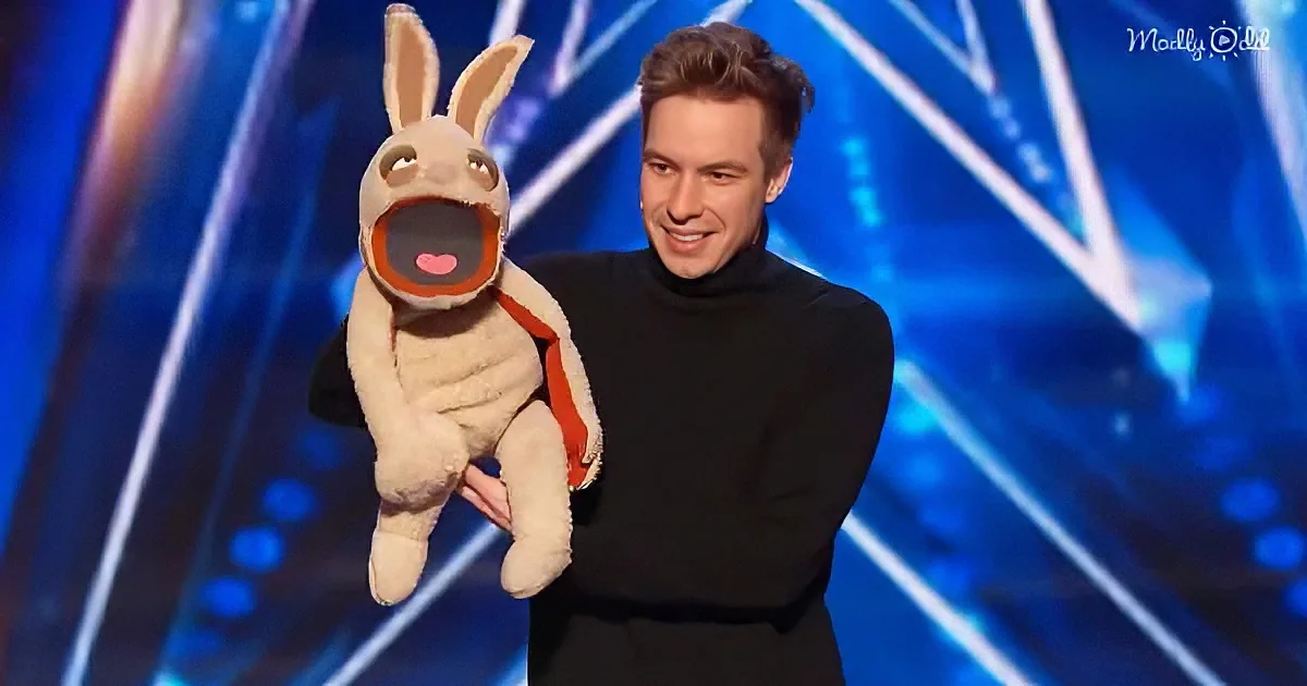 A man in a black turtleneck holds a puppet resembling a rabbit with a red scarf on a stage with blue lighting.
