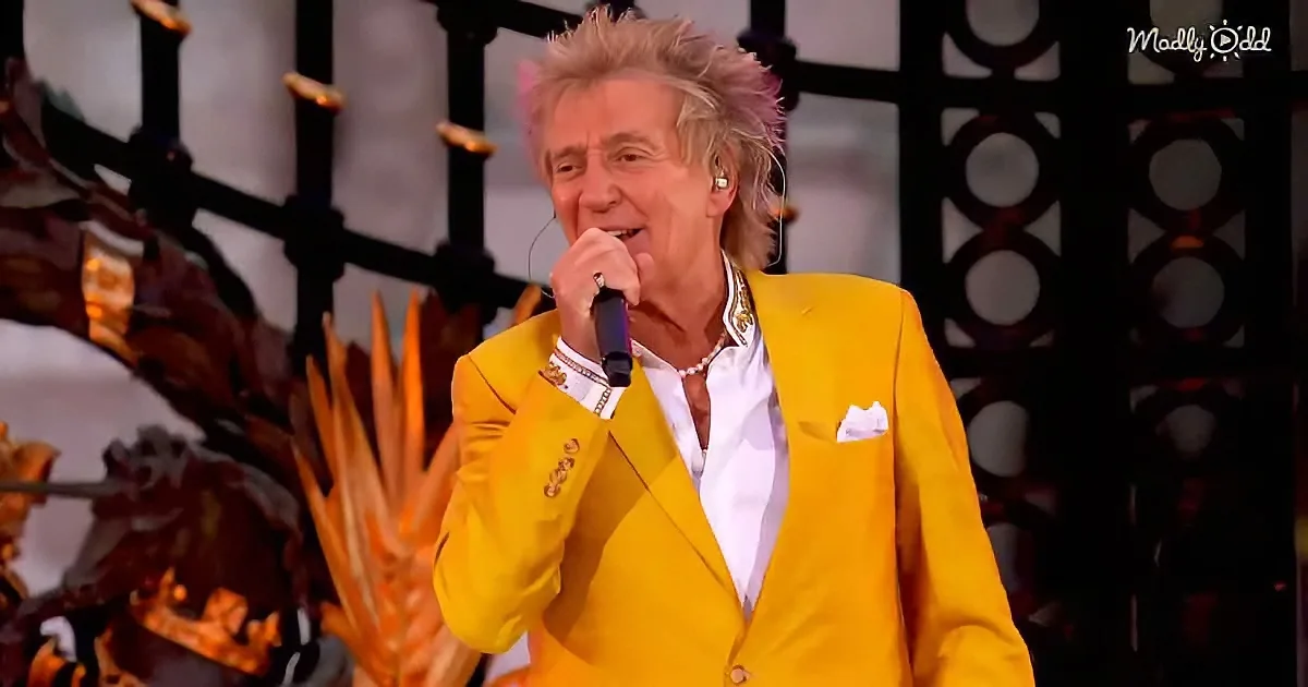 A person in a bright yellow suit sings into a microphone on stage, with a decorative background.
