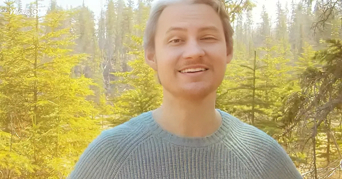 A man in a blue sweater smiles in front of a forest backdrop with sunlight filtering through the trees.
