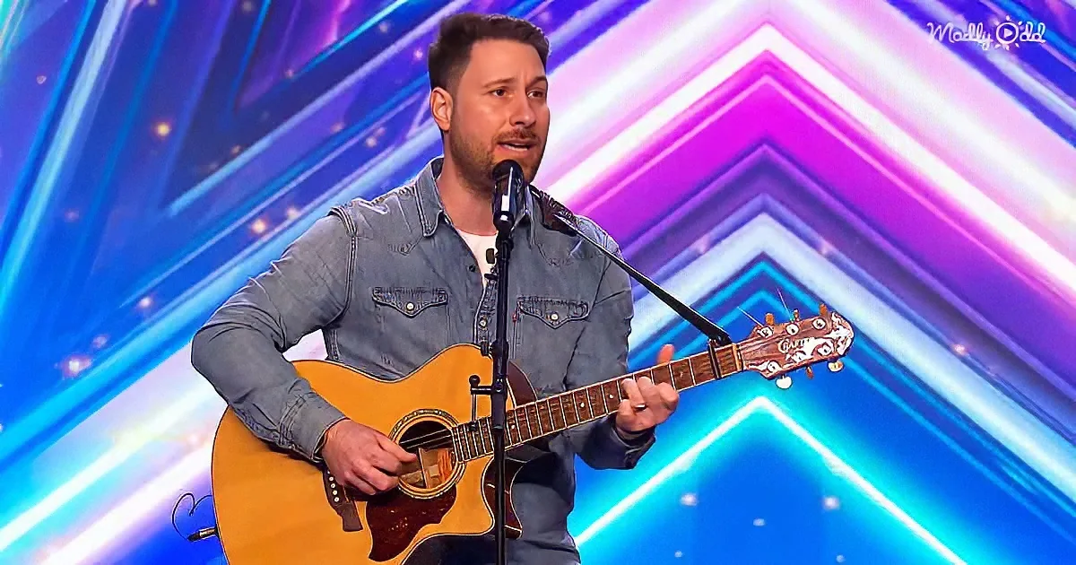 A man in a denim shirt plays an acoustic guitar on stage with a microphone.