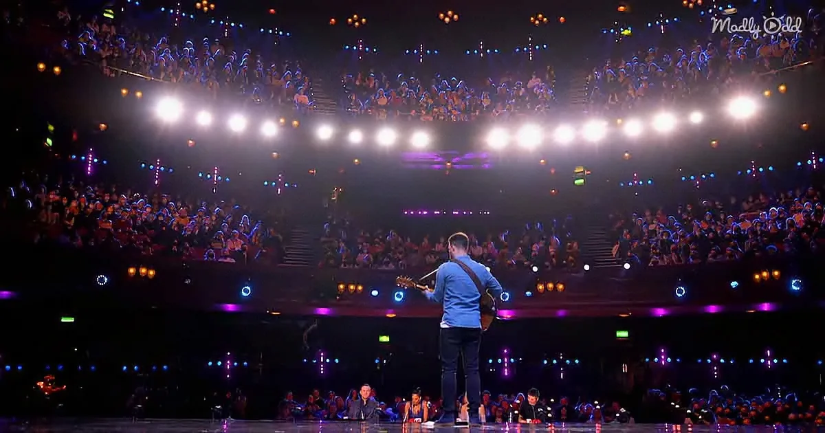 A man plays guitar on stage in front of a large, illuminated audience in a concert hall.