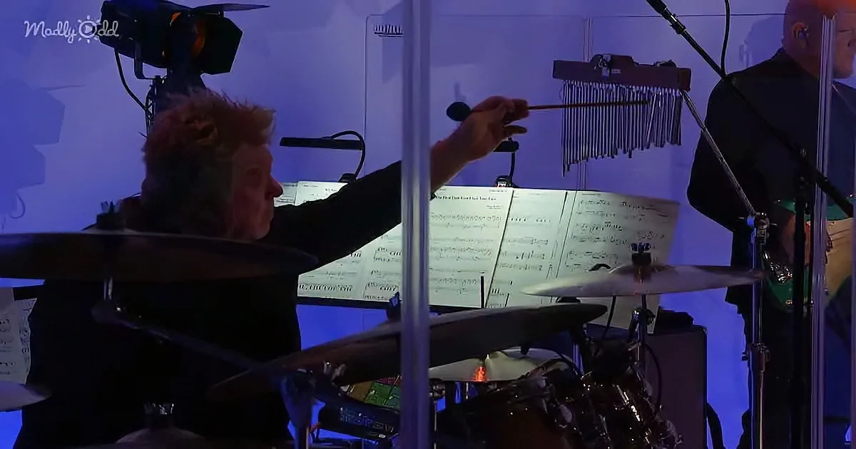 A musician plays a marimba with a mallet, surrounded by musical instruments and sheet music.