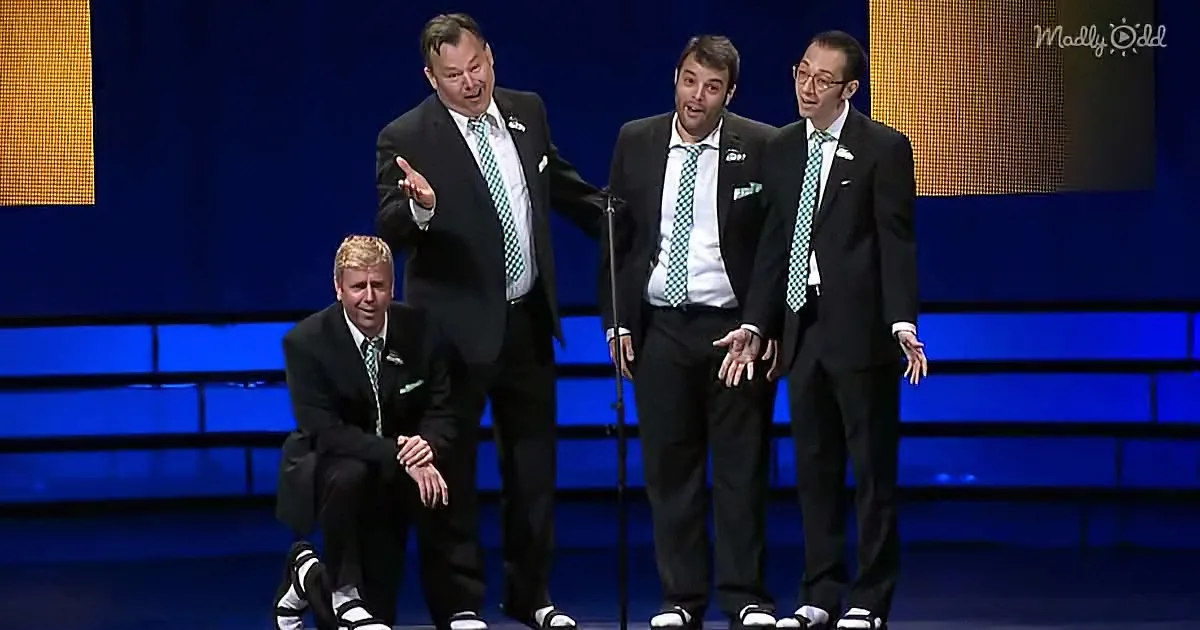Four men in suits and green ties perform on stage, one kneeling, others standing, with a microphone in front.