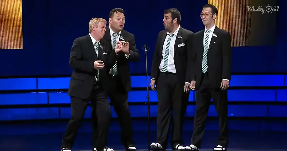 Four men in suits and green ties perform on stage, one holding a phone.