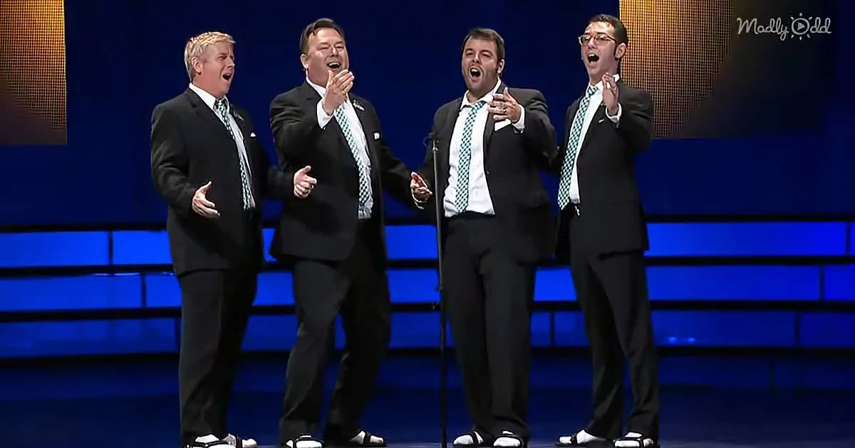 Four men in suits sing passionately on stage with a blue-lit background.