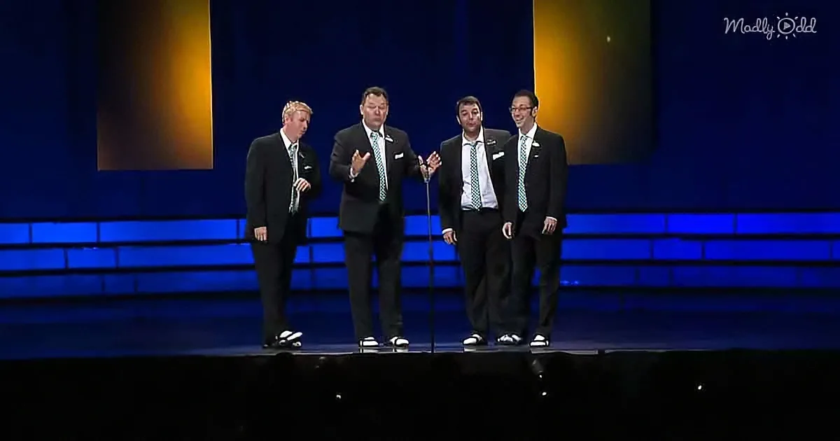 Four men in suits stand on stage, one gesturing, under blue and orange lighting.