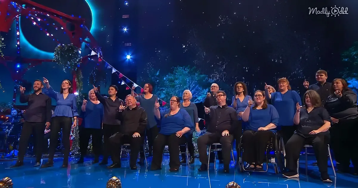 A group of people, some in wheelchairs, are on stage with a festive backdrop, pointing upwards.