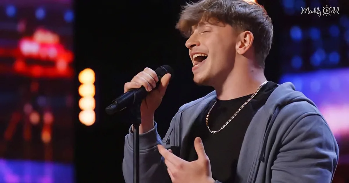 A person singing passionately into a microphone, wearing a black shirt and gold chain, with a colorful stage backdrop.