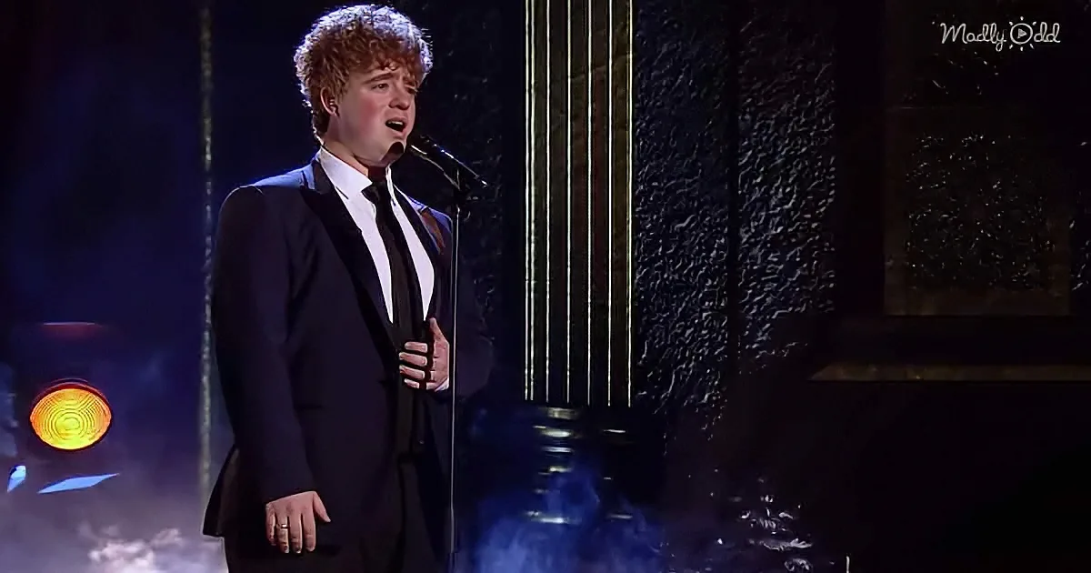A man in a black suit sings passionately on stage with a microphone, surrounded by dramatic lighting and smoke.