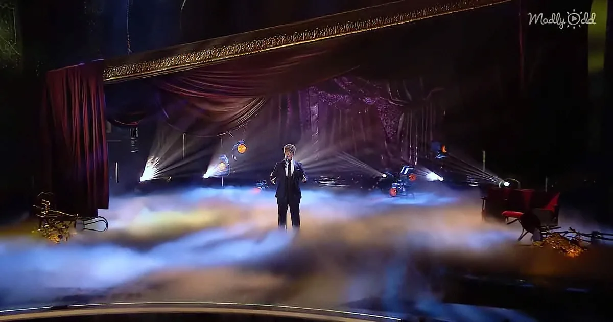 A man in a suit stands on a stage with dramatic lighting and fog, surrounded by red curtains and a grand backdrop.