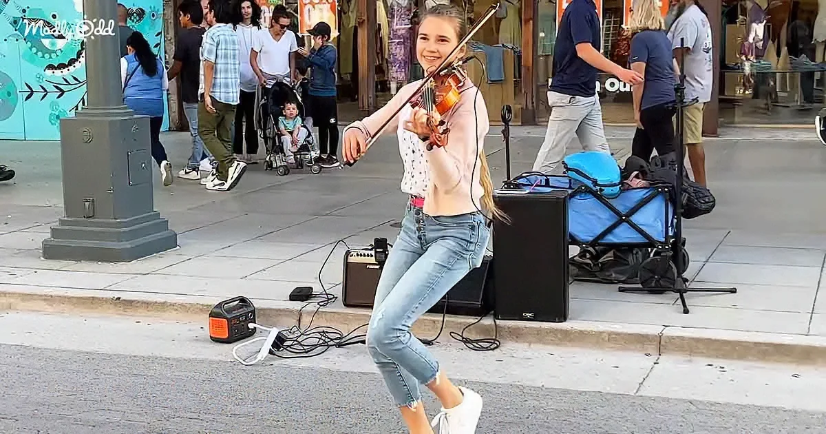A young woman plays violin on a city street, surrounded by pedestrians and musical equipment.