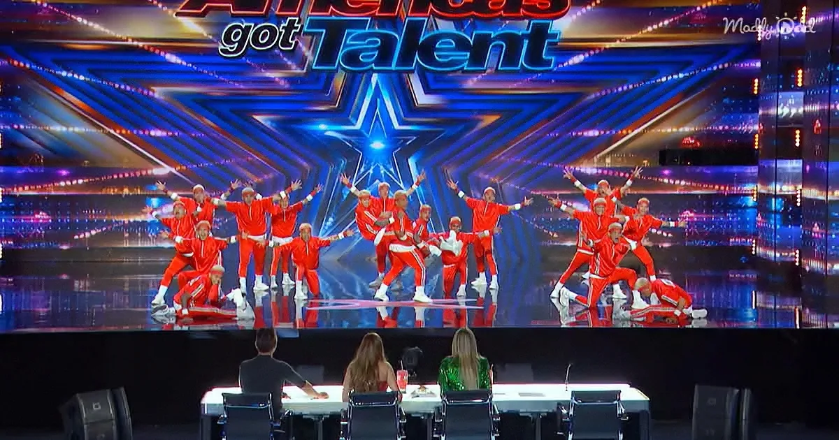 A group of dancers in red outfits performs on a stage with judges seated in front.