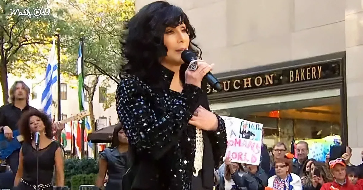 A person with long black hair and a black sequined outfit sings into a microphone in front of a crowd and flags.