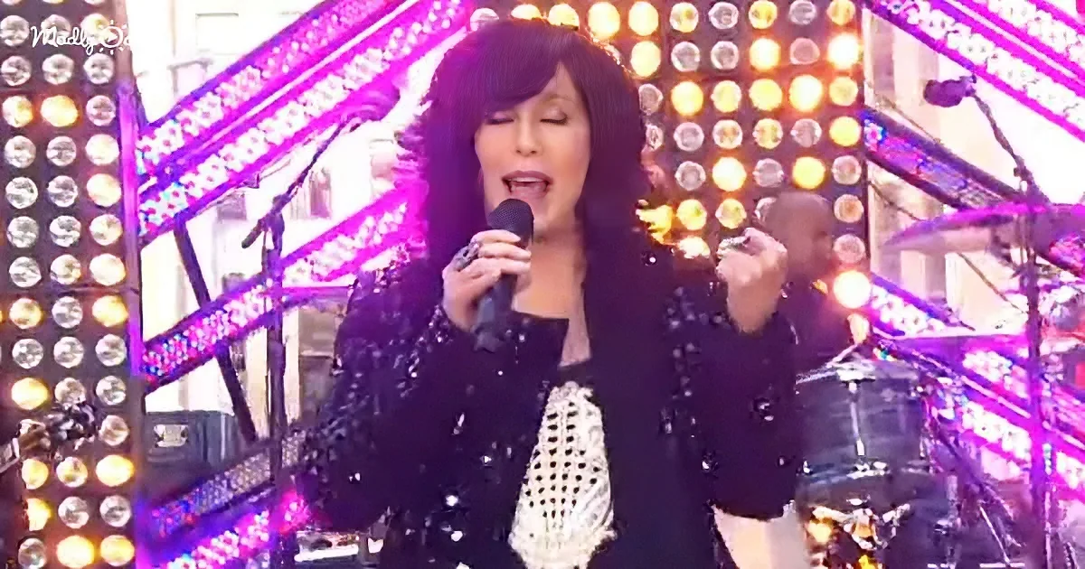 A woman in a black sequined outfit sings into a microphone on a stage with bright lights and a drummer in the background.