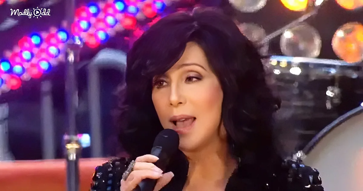 A woman with dark hair and a black outfit sings into a microphone, with colorful stage lights in the background.