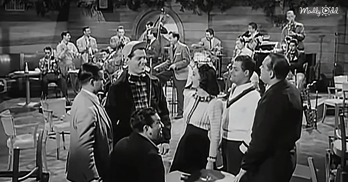 A man in a plaid shirt stands in a crowded room, surrounded by people and a band playing in the background.