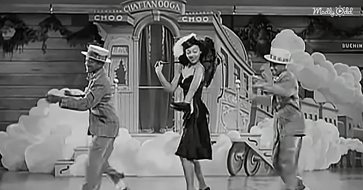 A woman in a black dress dances with two men in front of a train backdrop.
