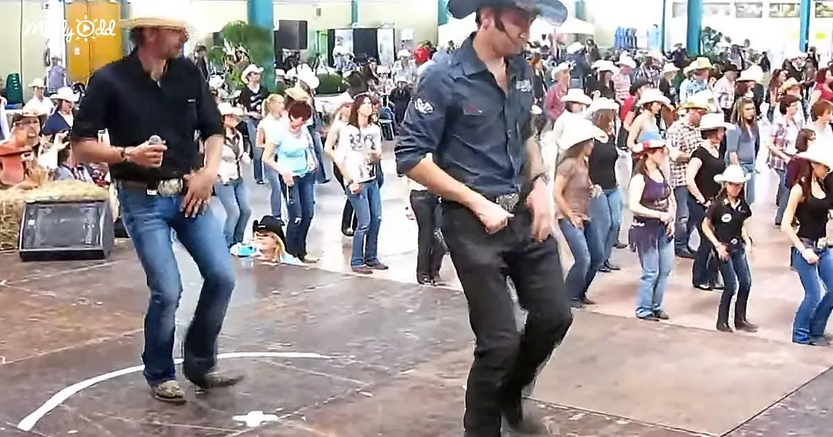 Two men in cowboy attire dance in a circle at a lively event with a crowd watching.