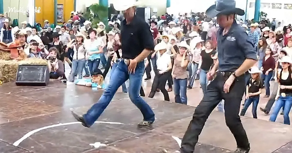 Two men in cowboy hats dance on stage, surrounded by a crowd of spectators at an outdoor event.
