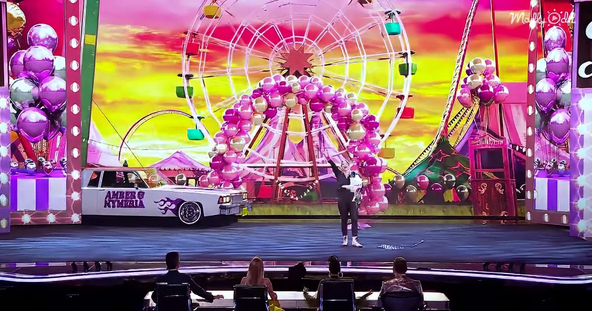 A person holds a dog on stage with a colorful ferris wheel and balloons in the background.