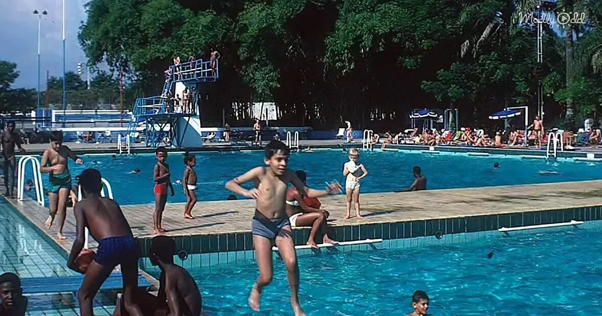 A boy jumps into a swimming pool surrounded by other children and adults enjoying the sunny day.