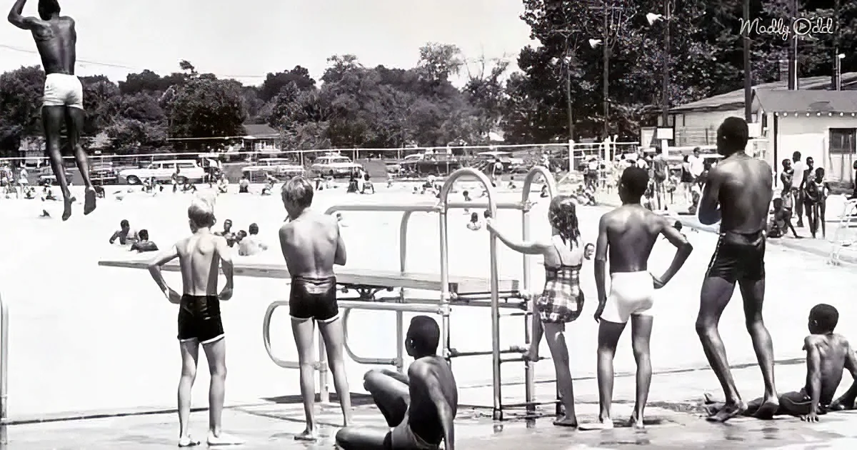 A man jumps off a diving board into a crowded swimming pool, surrounded by onlookers.