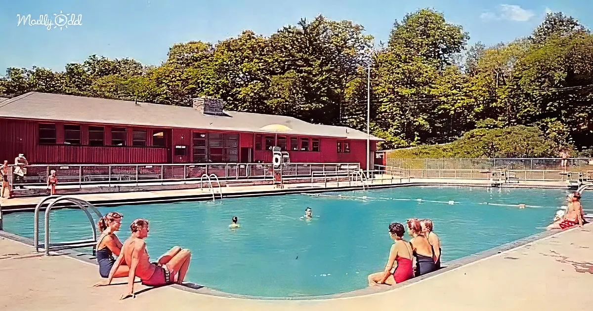 People relaxing and swimming in a large outdoor pool with a red building and trees in the background.